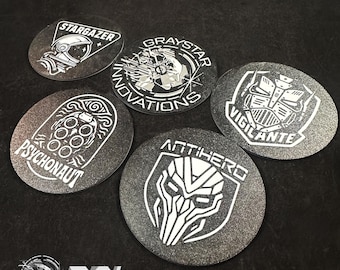 May include: A collection of five round, textured, dark gray metal coasters with white graphic designs and text. The designs include a space helmet, a robot, and the words "STARGAZER", "GRAYSTAR INNOVATIONS", "PSYCHONAUT", "ANTIHERO", and "VIGILANTE".