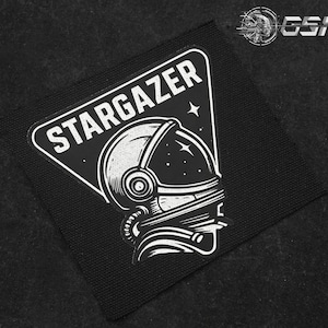 May include: Black and white patch featuring an astronaut helmet design with the word "STARGAZER" in a white, bold font. The patch has a triangular shape with rounded corners and a black background.