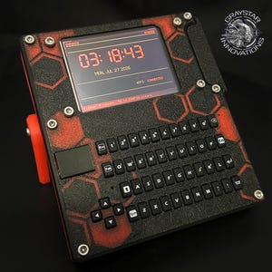 May include: A portable electronic device with a black and red hexagonal pattern, featuring a digital screen displaying the time 03:18:43, the date MON, JUL 27 2026, and WIFI: CONNECTED. It includes a QWERTY keyboard and a logo reading GRAYSTAR INNOVATIONS.