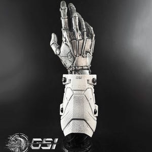Puede incluir: Una mano robótica plateada con intrincados detalles mecánicos. Está unida a un antebrazo blanco y blindado con el texto "GSI". La imagen se presenta sobre un fondo oscuro.