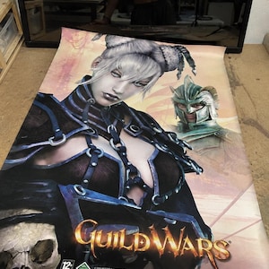 May include: A large Guild Wars promotional poster featuring a central character with white hair in buns, wearing dark armor with blue straps and chains. A second character in a green helmet appears behind. The Guild Wars logo is at the bottom.
