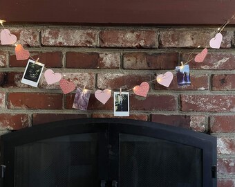 May include: A string of pink paper hearts with photos clipped to them hangs above a black fireplace. The hearts are strung with white lights.