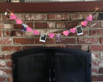 May include: A pink heart garland with photos clipped to each heart hangs above a black fireplace with a brick surround.