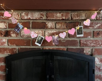 May include: A pink heart garland with string lights and photos clipped to it hangs above a black fireplace.