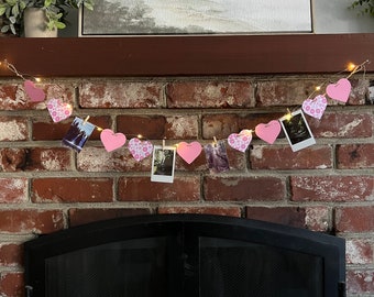 May include: A pink and white heart garland with photos clipped to it hangs above a black fireplace. The garland is strung with white fairy lights.