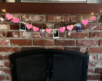 May include: A pink paper heart garland with string lights and instant photos clipped to each heart, hanging above a black fireplace with a brick surround.