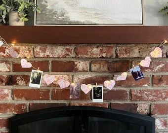 May include: A string of pink heart-shaped paper with photos hanging from it. The string is strung with small white lights and is hanging above a fireplace.