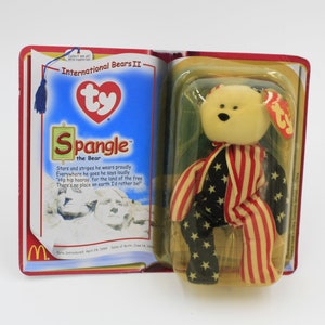 Puede incluir: Un oso de peluche Ty Beanie Baby llamado "Spangle" el oso, que lleva un atuendo rojo, blanco y azul con estrellas y rayas. El Beanie Baby está en su embalaje original, que presenta una imagen del Monte Rushmore.