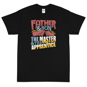 Op de afbeelding: Zwart T-shirt met een grafisch ontwerp met de tekst "Father & Son The Master & The Apprentice" met twee stotende vuisten.