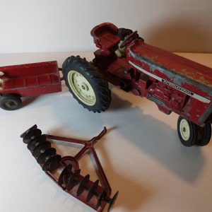May include: Red toy tractor with a wagon and a disc harrow. The tractor has the words "International" on the side.