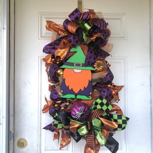 Könnte beinhalten: Ein Halloween-Kranz mit einem Zwerg, der einen grünen Hut und einen orangefarbenen Bart trägt. Der Kranz ist mit orangefarbenen, grünen und violetten Bändern sowie schwarz-weißem kariertem Stoff verziert.