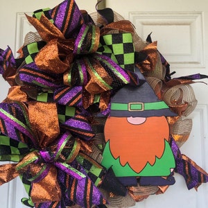 May include: A Halloween wreath featuring orange, green, black and purple ribbon, burlap and mesh. A gnome with a green hat and orange beard is in the centre.