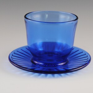 May include: A cobalt blue glass cup and saucer set. The cup has a rounded shape and the saucer has a fluted edge.