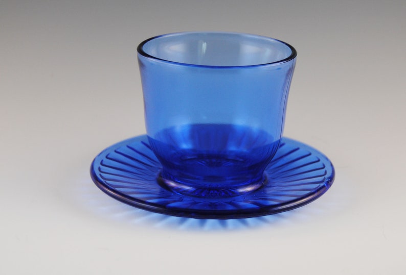 May include: A cobalt blue glass cup and saucer set. The cup has a rounded shape and the saucer has a fluted edge.