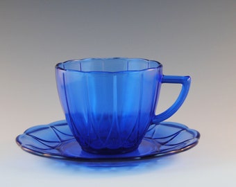 May include: A cobalt blue glass teacup and saucer set. The cup has a handle and vertical ribbed detailing. The saucer has a scalloped edge. The set is made of transparent glass.