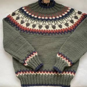 Könnte beinhalten: Olivgrüner Strickpullover mit hohem Kragen und langen Ärmeln. Der Halsausschnitt hat ein sich wiederholendes Muster in Weiß, Braun, Rot und Marineblau. Die Manschetten und der Saum haben ein marineblaues, weißes und rotes Zickzackmuster.