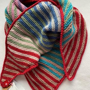 Puede incluir: Una bufanda triangular tejida a mano con un vibrante patrón de rayas en tonos rojo, azul, verde, rosa y beige. La bufanda tiene un borde de ganchillo rojo distintivo e incluye una etiqueta blanca de producto DROPS MERINO EXTRA FINE.