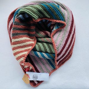 Puede incluir: Una bufanda infinita tejida a mano con rayas horizontales en tonos rosa, burdeos, verde oliva, azul cerceta y crema. El tejido tiene una textura de punto bobo e incluye una etiqueta blanca con el texto DROPS MERINO EXTRA FINE.