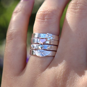 May include: A hand wearing a stack of five silver rings with different charms. The charms include a flower, a heart, a star, and a leaf.