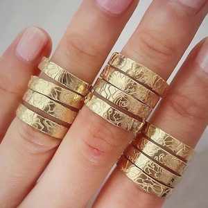 May include: A hand wearing a stack of gold rings with a floral design. The rings are stacked on the middle finger.