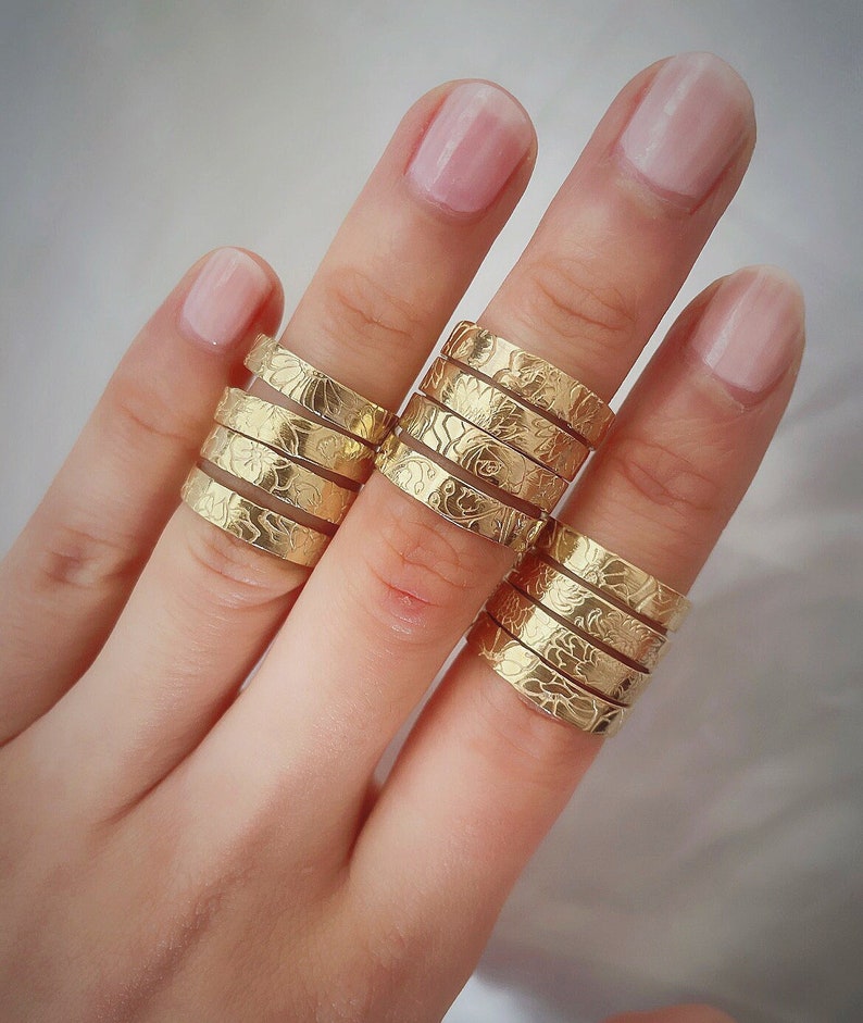 May include: A hand wearing a stack of gold rings with a floral design. The rings are stacked on the middle finger.