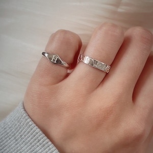 May include: Two silver rings on a finger. One ring is a simple band with a textured surface. The other ring is a wider band with a raised letter "H" on a oval-shaped surface.