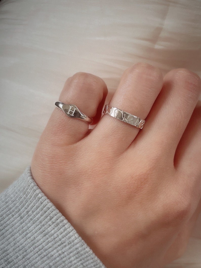 May include: Two silver rings on a finger. One ring is a simple band with a textured surface. The other ring is a wider band with a raised letter "H" on a oval-shaped surface.