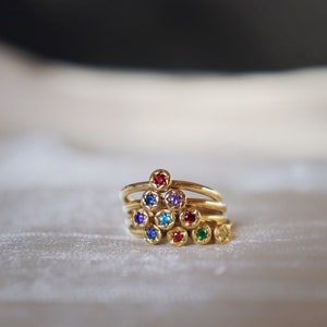 Puede incluir: Una pila de anillos dorados, cada uno adornado con una piedra preciosa de diferente color. Las piedras preciosas incluyen rojo, azul, morado, turquesa y verde. Los anillos están apilados en forma de pirámide.