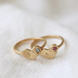 Puede incluir: Dos anillos dorados con caras en forma de corazón. Un anillo tiene la letra "S" grabada en el corazón y una pequeña gema azul. El otro anillo tiene la letra "A" y una pequeña gema roja.