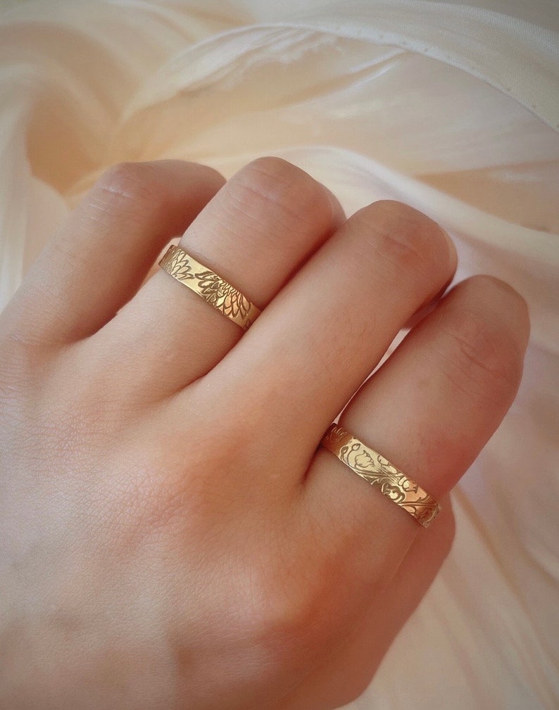 May include: Two gold rings with a floral design. The rings are worn on the fingers of a hand.