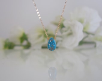 Puede incluir: Collar con cadena dorada y colgante en forma de lágrima turquesa con vetas marrones. El collar se exhibe sobre un fondo borroso de flores blancas. Un diseño delicado y elegante.