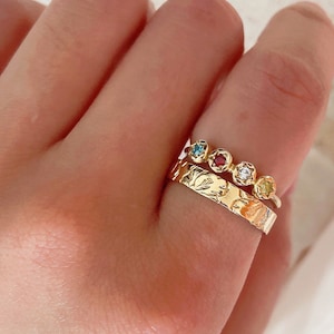 May include: A gold ring with a textured band and a row of five colorful gemstones. The gemstones are blue, red, clear, and yellow.
