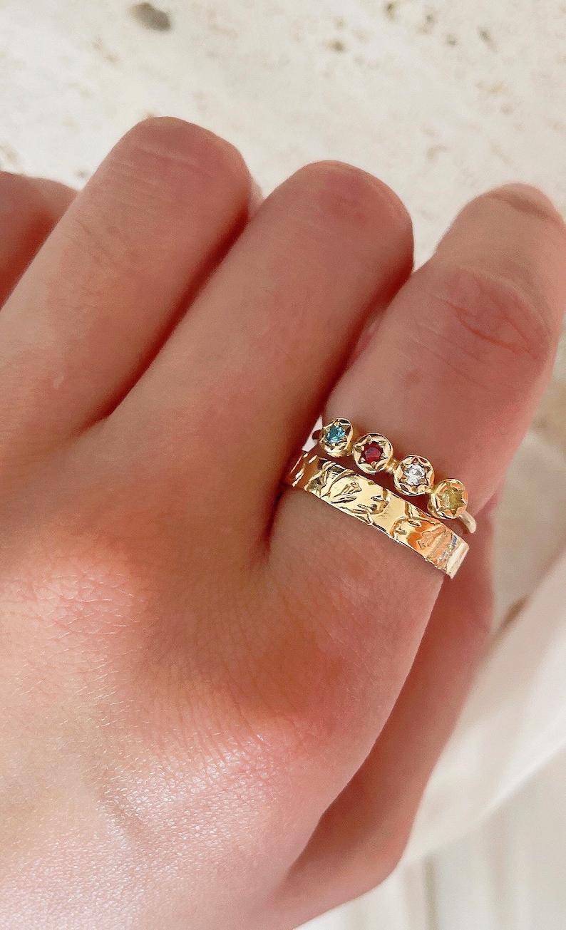 May include: A gold ring with a textured band and a row of five colorful gemstones. The gemstones are blue, red, clear, and yellow.