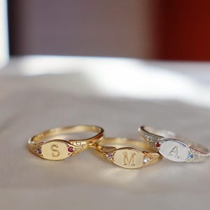 Puede incluir: Tres anillos de metal con placas frontales ovaladas grabadas con las letras S, M y A. Dos anillos son de tono dorado y uno de tono plateado. Cada anillo tiene filigranas en la banda y está decorado con pequeñas piedras preciosas de colores.