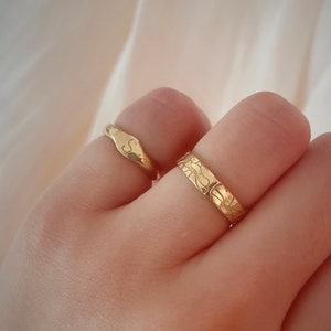 May include: Two gold rings on a finger. The ring on the index finger has a textured design. The ring on the middle finger is a simple band with an engraved letter 'S'.