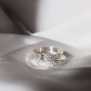 May include: Two silver rings with intricate floral designs. The rings are stacked on top of each other and are lying on a white satin fabric.