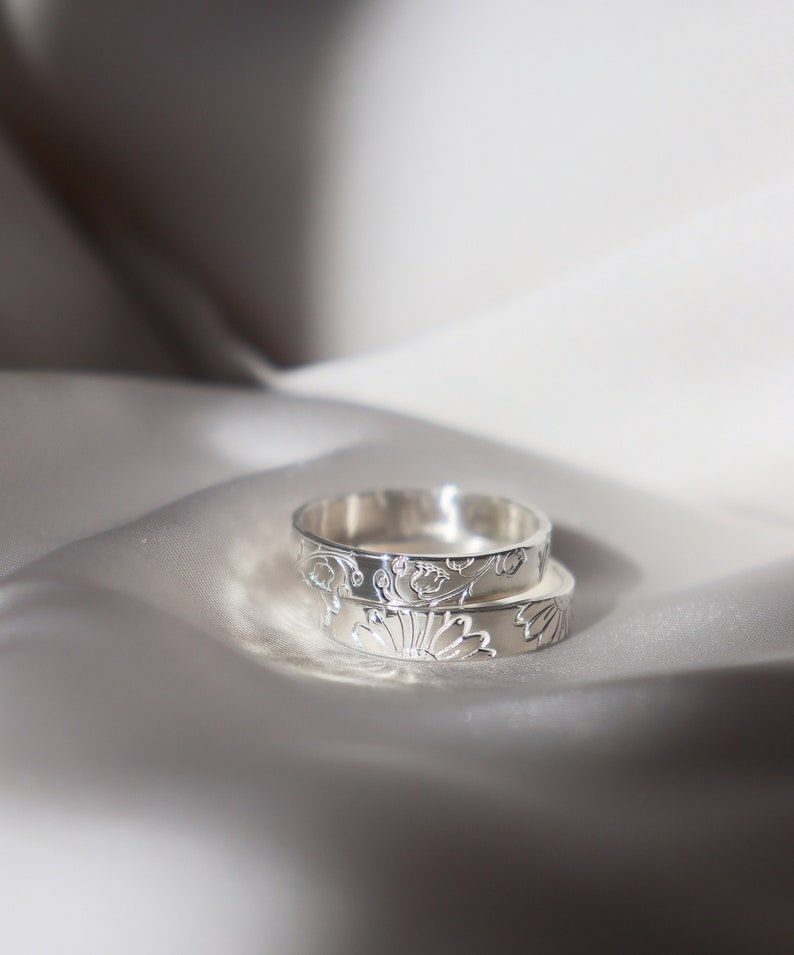 May include: Two silver rings with intricate floral designs. The rings are stacked on top of each other and are lying on a white satin fabric.