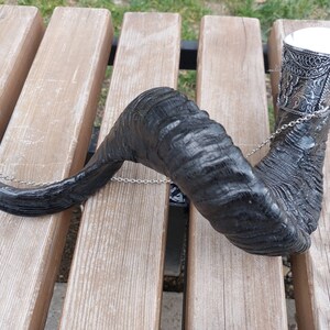 Op de afbeelding: Een zwart, hoornvormig drinkvat met sierlijke zilverkleurige metalen details. Een zilveren ketting verbindt de beker met een decoratief zilverkleurig metalen stuk. De hoorn rust op een houten bank.