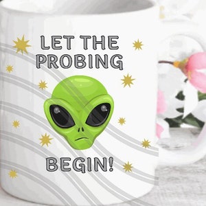 May include: White ceramic mug featuring a green alien face and the text "Let the Probing Begin!" in black lettering. The background is a white and grey striped pattern with small yellow stars.