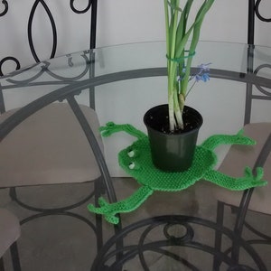 May include: A green crocheted frog-shaped coaster with a black pot of yellow daffodils in the center. The coaster is on a glass table with two black metal chairs.