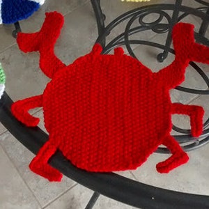 May include: Red crocheted crab-shaped trivet with a textured surface. The trivet is sitting on a glass table with two crocheted beach ball trivets.