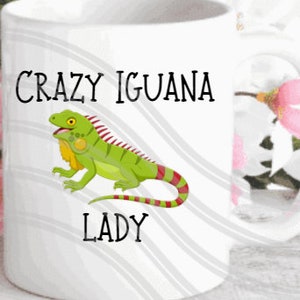 Könnte beinhalten: Weiße Keramik-Tasse mit einer Illustration einer grünen Leguan-Eidechse mit roten und gelben Akzenten. Der Text "CRAZY IGUANA LADY" ist in Schwarz gedruckt. Die Tasse hat einen Standardgriff und ist für heiße oder kalte Getränke geeignet.