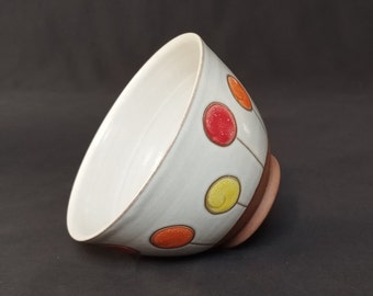 May include: A white ceramic bowl with a light blue glaze and a brown rim. The bowl is decorated with colorful circles in shades of red, orange, and yellow.