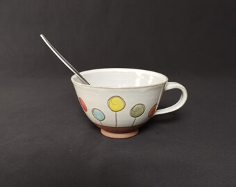 May include: A white ceramic teacup with a handle and a decorative pattern of colorful circles resembling flowers. A silver spoon rests inside the cup. The cup sits on a brown base.