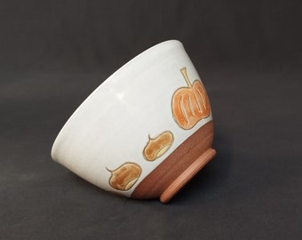 May include: A white ceramic bowl with a brown rim and a design of pumpkins and acorns. The bowl is tilted to the side.