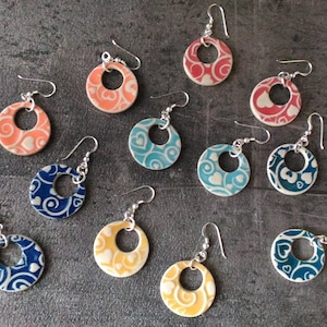 Puede incluir: Surtido de pendientes de cerámica con una variedad de patrones coloridos. Cada pendiente presenta un diseño circular con un patrón de corazón y un gancho plateado.