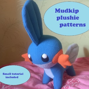 Pode incluir: Pelúcia Mudkip azul e laranja com um pequeno tutorial incluído. A pelúcia está sobre uma superfície rosa com o texto "Mudkip plushie patterns" em um círculo azul.