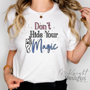 Op de afbeelding: Wit T-shirt met de tekst "Don't Hide Your Magic" in roze en blauw. Het ontwerp bevat een handgebaar en sparkles. Het T-shirt heeft een casual stijl.