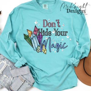 Könnte beinhalten: Ein türkisfarbenes Langarmshirt mit dem Text "Don't Hide Your Magic" und einer Grafik von bunten Kristallen. Das Design umfasst rosa, lila, grüne, blaue und gelbe Kristalle mit funkelnden Akzenten. Das Shirt ist von der Marke Comfort Colors.