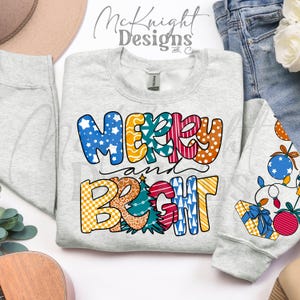 May include: A light gray sweatshirt with the words "MERRY and BRIGHT" in colorful, patterned letters. The design includes stars, polka dots, stripes, and a Christmas tree. The sleeve of the sweatshirt has a Christmas ornament design.